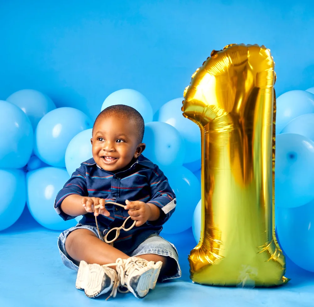 Baby in a blue shirt beside a gold birthday balloon